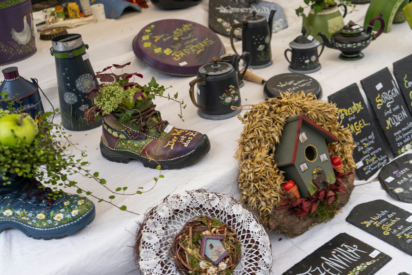Artesanía hecha a mano, como macetas pintadas con forma de bota y pizarras decoradas, en exhibición.